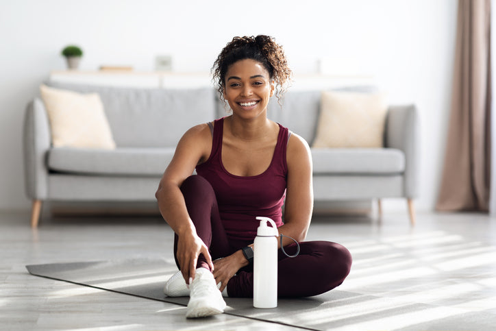 Eine junge Frau in sportlicher Kleidung sitzt lächelnd auf einer Yogamatte in einem hellen Wohnzimmer. Neben ihr steht eine Wasserflasche – bereit für das nächste Workout oder einen Moment der Erholung.